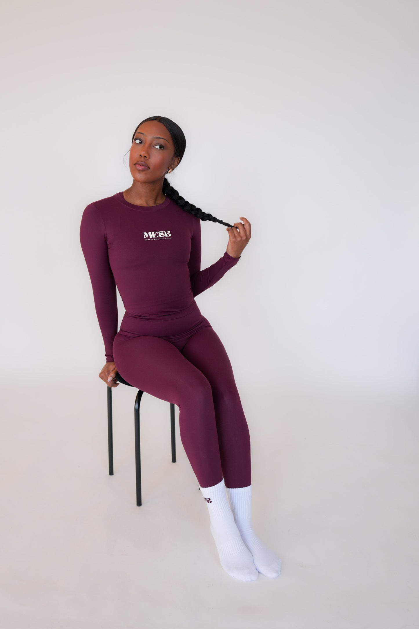 Model sitting on black stool with white background wearing the long sleeve Garnet glow set from MESB. She is playing with her hair and looking to the side